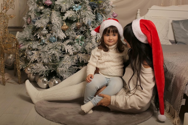 Mädchen in Pullover und Kniestrümpfen sitzt am Bett am Weihnachtsbaum und umarmt ihre Tochter