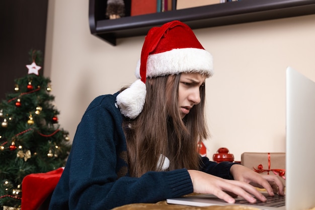 Mädchen in Santa Claus Hut spricht mit Laptop für Videoanruf Freunde und Eltern. Das Zimmer ist festlich eingerichtet.