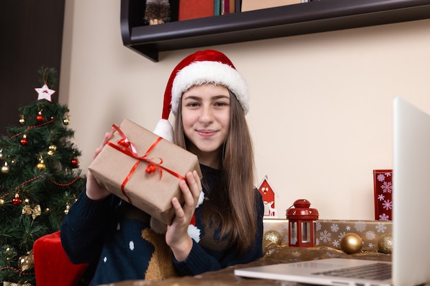 Mädchen in Santa Claus Hut spricht und gibt ein Geschenk mit Laptop für Videoanruf Freunde und Eltern. Das Zimmer ist festlich eingerichtet. Weihnachten während des Coronavirus.