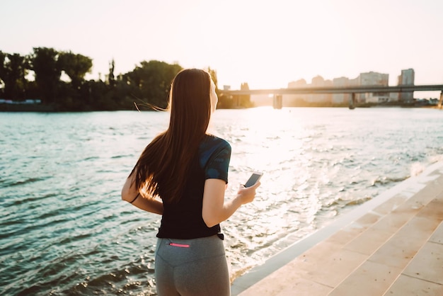 Mädchen in Sportuniform mit Kopfhörern läuft bei Sonnenuntergang am Fluss entlang