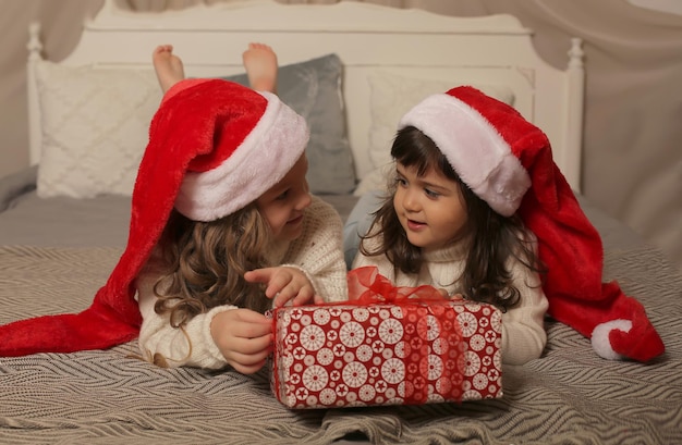 Mädchen in Weihnachtsmannmützen und weißen Pullovern liegen mit einer Geschenkbox in der Hand auf dem Bett