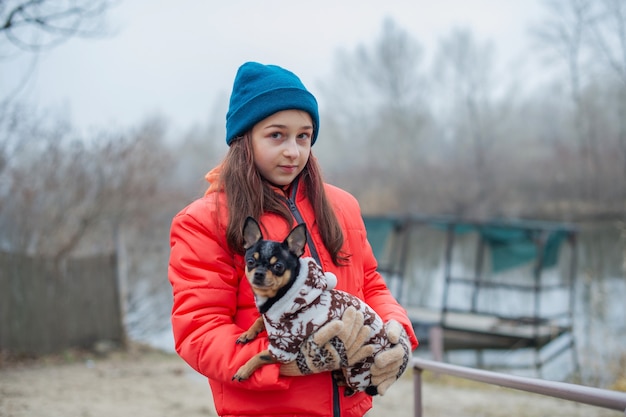 Mädchen in winterkleidung. teenager-mädchen in einer orange jacke, mütze und schal. mädchen und chihuahua.