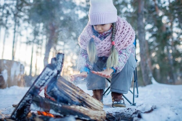 Mädchen in Winterkleidung wärmt sich am Wochenende am Feuer im verschneiten Wald auf