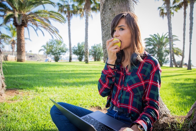 Mädchen isst einen Apfel, während sie den Laptop in einem Park im Freien benutzt