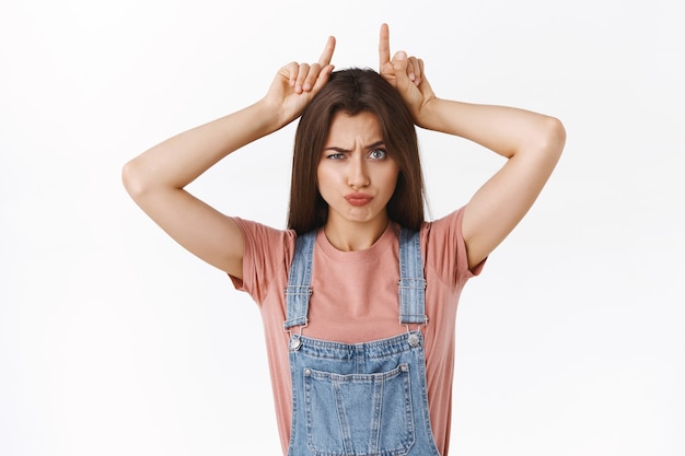 Mädchen ist stur und wird niemals aufgeben. Verspielte süße und lustige brünette Frau in Overalls, T-Shirt, mit Stierhörnern und Grimassen, wie sie entschlossen sind zu gewinnen, stehender weißer Hintergrund