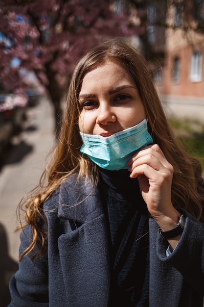 Mädchen, junge Frau in einer sterilen medizinischen Schutzmaske auf ihrem Gesicht, die die Kamera im Freien betrachtet, im Frühlingsgarten. Luftverschmutzung, Virus, pandemisches Coronavirus-Konzept.