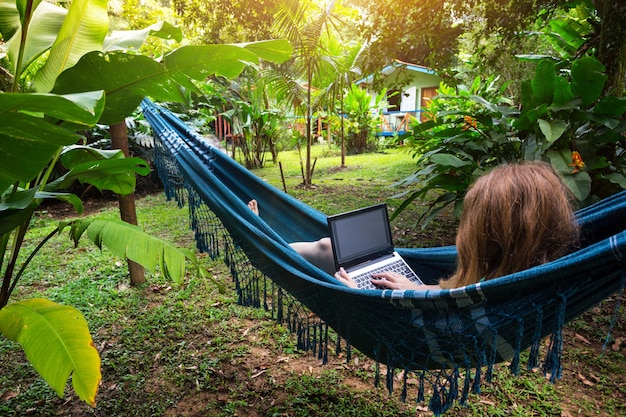 Mädchen liegt in einer Hängematte und arbeitet im Garten an einem Laptop