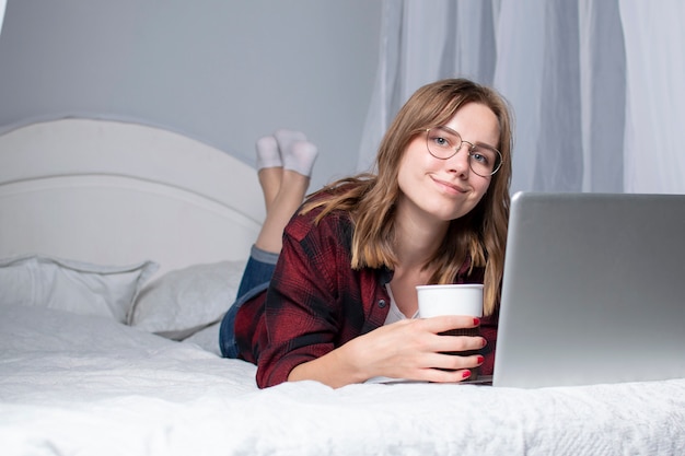 Mädchen liegt mit einem Laptop im Bett und schaut sich ein Video mit Kaffee an