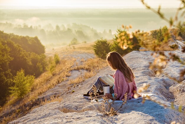 Mädchen liest Buch, während es gegen schöne Naturlandschaft sitzt. Sie hält Buch in Bergen. Sommerurlaub und Lifestyle-Konzept