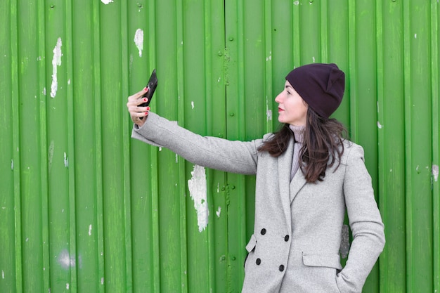 Mädchen macht ein Selfie an einer grünen Industrietür. Hut und Mantel tragen. Platz zum Schreiben. gekleidet in eine kalte Pagode. Kleidung und Stil. natürliche Emotionen