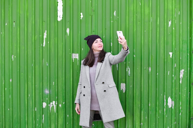 Mädchen macht ein Selfie an einer grünen Industrietür. Hut und Mantel tragen. Platz zum Schreiben. gekleidet in eine kalte Pagode. Kleidung und Stil. natürliche Emotionen