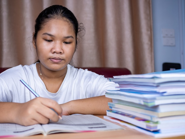 Mädchen macht Hausaufgaben auf einem Holztisch und daneben lag ein Stapel Bücher Der Hintergrund ist ein rotes Sofa und cremefarbene Vorhänge.