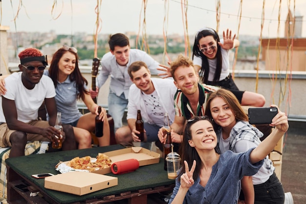 Mädchen macht Selfie mit leckerer Pizza Eine Gruppe junger Leute in legerer Kleidung feiern tagsüber gemeinsam auf dem Dach