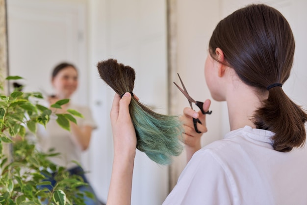 Mädchen macht zu Hause einen Haarschnitt, Teenager schneidet sich die Haare, in den Händen ein Bündel geschnittener Haare und eine Schere, schneidet gefärbtes ungesundes Haar