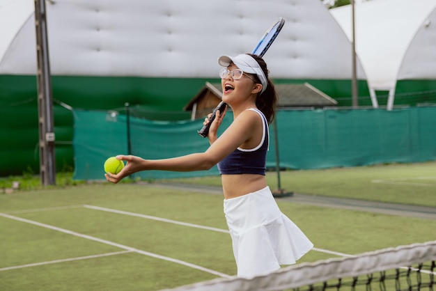 Mädchen mit asiatischem Aussehen in Brille und Mütze spielt Tennis auf dem Tennisplatz