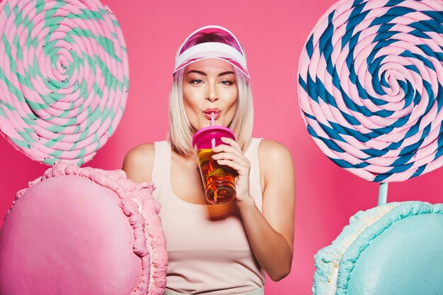 Mädchen mit blondem Haar, das Spitze und rosa Kappe trägt, die mit riesigen süßen Lutschbonbons an Rosa stehen