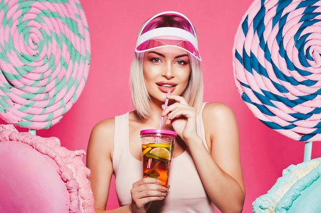 Mädchen mit blondem Haar, das Spitze und rosa Kappe trägt, die mit riesigen süßen Lutschbonbons an Rosa stehen