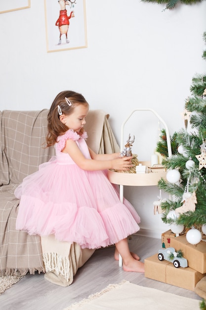Mädchen mit blonden Haaren in einem schicken rosa Kleid neben dem Weihnachtsbaum sitzt auf dem Sofa und spielt