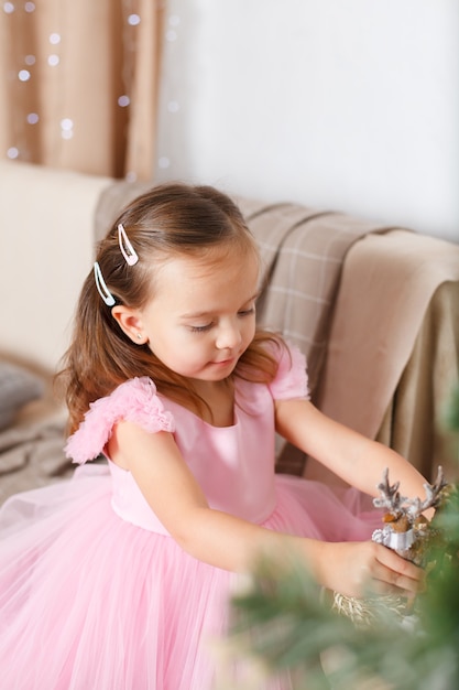 Mädchen mit blonden Haaren in einem schicken rosa Kleid neben dem Weihnachtsbaum sitzt auf dem Sofa und spielt