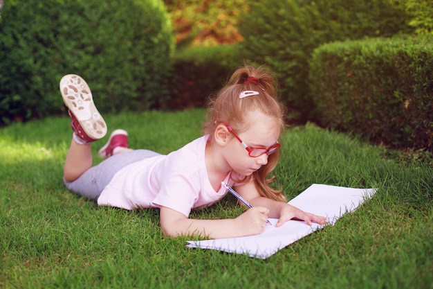 Mädchen mit Brille liegt im Gras und zeichnet auf Papier