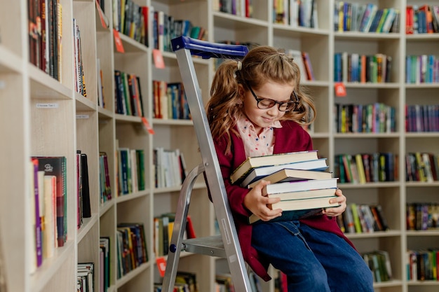 Mädchen mit Brille liest ein Buch, das auf einer Trittleiter in der Bibliothek sitzt