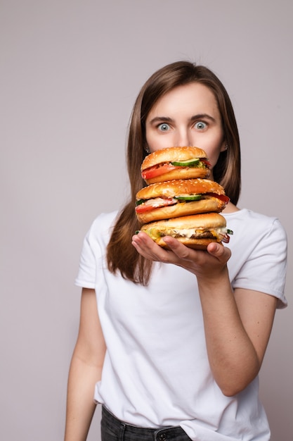 Mädchen mit dem riesigen Hamburger an der Hand. Studio-Porträt der jungen brünetten Frau im weißen T-Shirt, das enorme Burger auf ihrer Hand hält, die schockiert oder in die Kamera überrascht schaut.