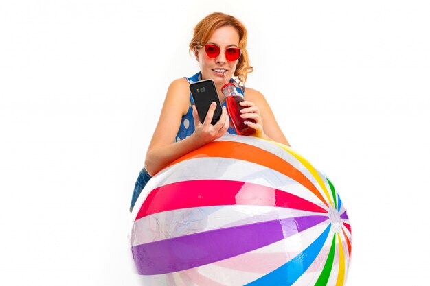 Mädchen mit einem aufblasbaren Ball zum Schwimmen, einem Telefon und einem Glas Cocktail in ihren Händen auf einer weißen Wand