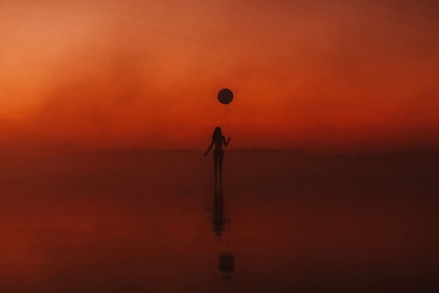 Mädchen mit einem Ballon in ihrer Hand auf dem Wasser bei Sonnenaufgang im Nebel im Sommer