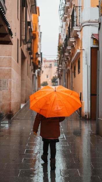 Mädchen mit einem orangefarbenen Regenschirm, der sich vor dem Regen schützt, der eine einsame schmale Straße hinuntergeht