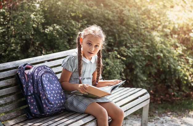 Mädchen mit einem Rucksack, der auf einer Bank sitzt und ein Buch in der Nähe der Schule liest Zurück zum Unterrichtsplan der Schule ein Tagebuch mit Noten Bildung Grundschulklassen Zurück zur Schule