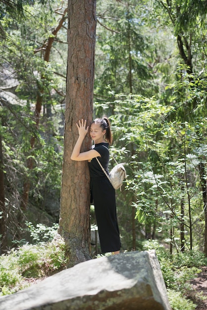 Mädchen mit einem Rucksack umarmt einen Baum mit geschlossenen Augen im Wald. Sonniger Sommertag.