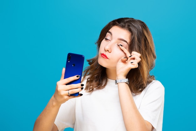 Mädchen mit einem Telefon in ihren Händen lächelt und macht Make-up, isoliert auf einem Blau.