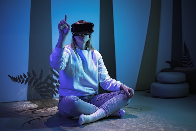 Mädchen mit einem virtuellen Realitäts-Headset sitzt in einer Lotus-Pose und meditiert in einem Raum mit blauem Licht