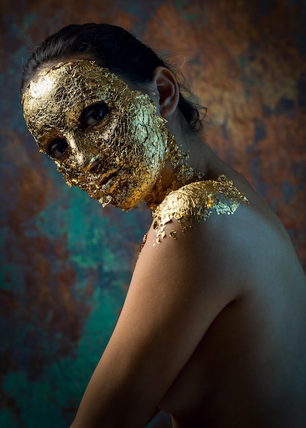 Mädchen mit einer Maske auf ihrem Gesicht aus Blattgold Düsteres Studioporträt einer Brünette auf abstraktem Hintergrund