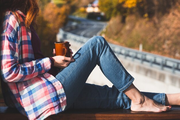Mädchen mit einer Tasse Kaffee, die auf dem Geländer auf dem Balkon sitzt. Morgen in einer Berghütte
