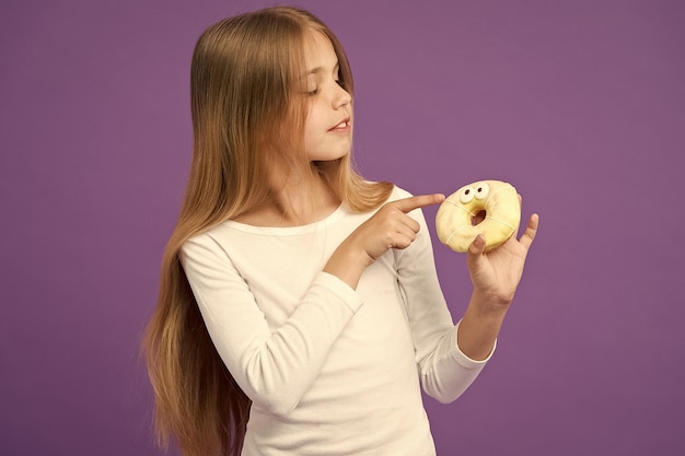 Mädchen mit flauschigen Donut Glasierter Bagel mit Augen kreatives Dessert Kind isst Vereisung aus Donut-Naschkatzenhimmel Kind mit langen Haaren, die einen weißen Pullover tragen, der auf violettem Hintergrund isoliert ist