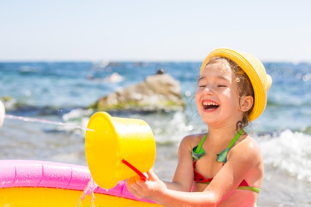 Mädchen mit gelbem Strohhut spielt mit dem Windwasser und einem Wasserspender in einem aufblasbaren Pool am Strand. Unauslöschliche Produkte zum Schutz der Kinderhaut vor Sonnenbrand am Meer