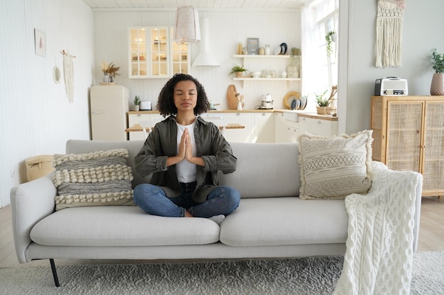 Mädchen mit gemischten Rassen praktiziert Yoga-Meditation zu Hause und sitzt in Lotus-Pose auf der Couch Gesunder Lebensstil