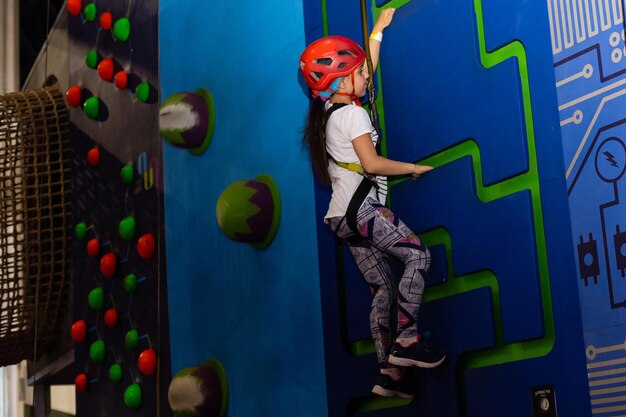 Mädchen mit Gurt und Sicherheitsausrüstung klettert an praktischer Wand im Innenbereich. Mädchen führt das Seil in eine Expressschlinge ein.