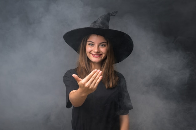 Mädchen mit Hexenkostüm für Halloween-Partys über der lokalisierten dunklen Wand, die einlädt zu kommen