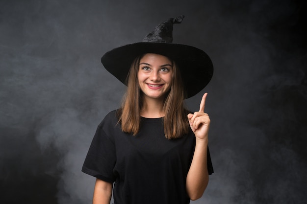 Mädchen mit Hexenkostüm für Halloween-Partys über der lokalisierten dunklen Wand, die mit dem Zeigefinger eine großartige Idee zeigt