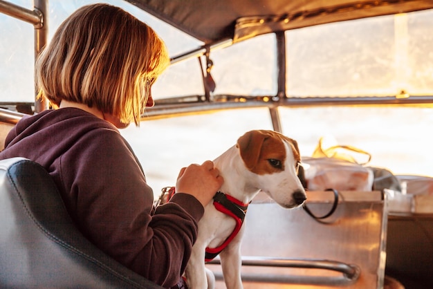 Mädchen mit Hund Jack Russell Terrier in ihren Armen, das bei Sonnenuntergang in einem Boot am Meer sitzt