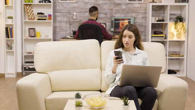 Mädchen mit ihrem Telefon und Laptop beim Sitzen auf der Couch. Freund im Hintergrund.