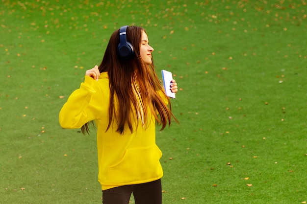 Mädchen mit Kopfhörern auf einer grünen Wiese in einem gelben Hoodie tanzen, Musik hören