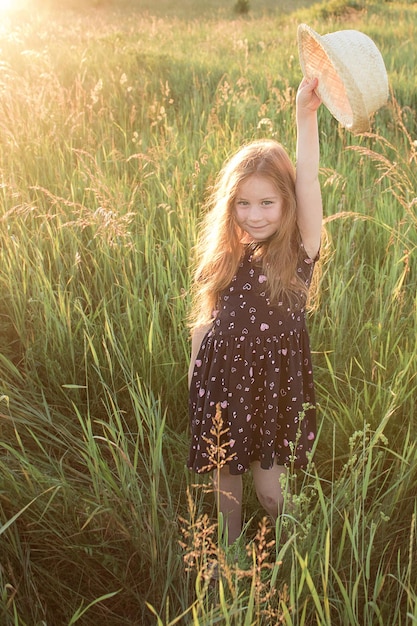 Mädchen mit langen Haaren steht im Sommer im hohen Gras und hält einen Strohhut in der erhobenen Hand