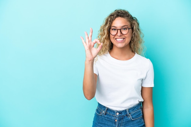 Mädchen mit lockigem Haar auf blauem Hintergrund isoliert, das mit den Fingern ein OK-Zeichen zeigt