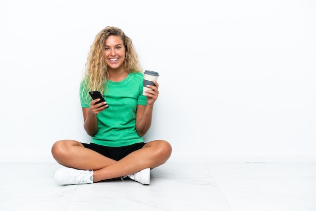 Mädchen mit lockigem Haar, das auf dem Boden sitzt und Kaffee zum Mitnehmen und ein Handy hält