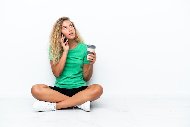 Mädchen mit lockigem Haar, das auf dem Boden sitzt und Kaffee zum Mitnehmen und ein Handy hält