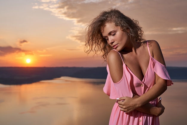 Mädchen mit lockigem Haar im rosa Kleid auf Hintergrund des Sonnenuntergangs.