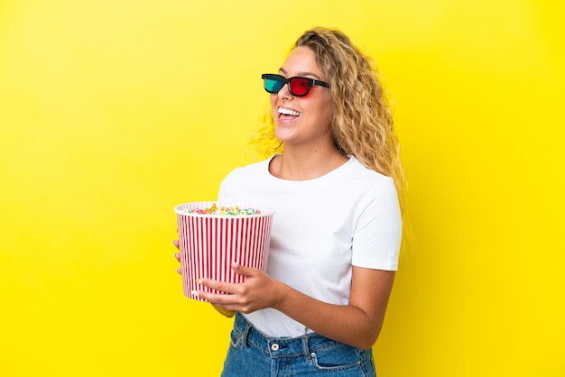 Mädchen mit lockigem Haar isoliert auf gelbem Hintergrund mit 3D-Brille und mit einem großen Eimer Popcorn